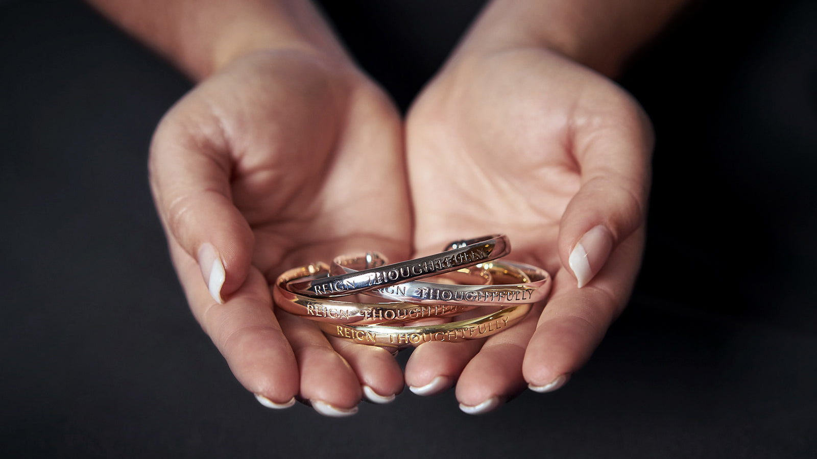Woman holding stack of engraved "Reign Thoughtfully" Cuffs from REALM in gold, silver, rose gold and black ruthenium.
