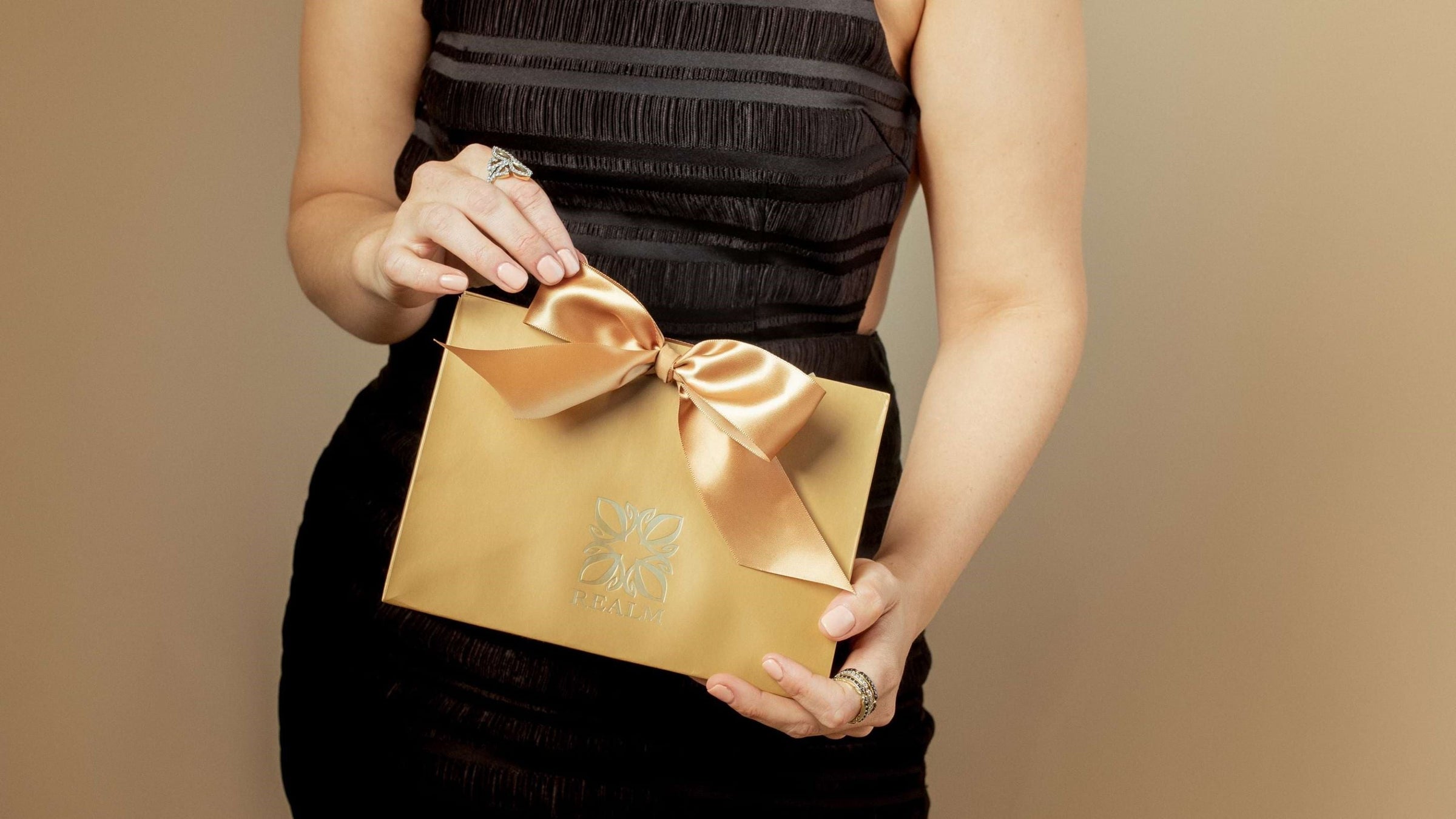 woman in black dress holds luxe REALM Fine + Fashion Jewelry gold gift bag