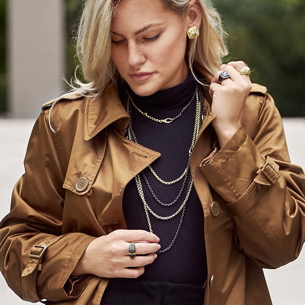 Young woman wearing layered REALM Everyday Chain necklaces in mixed metals with a brown trench coat and black top